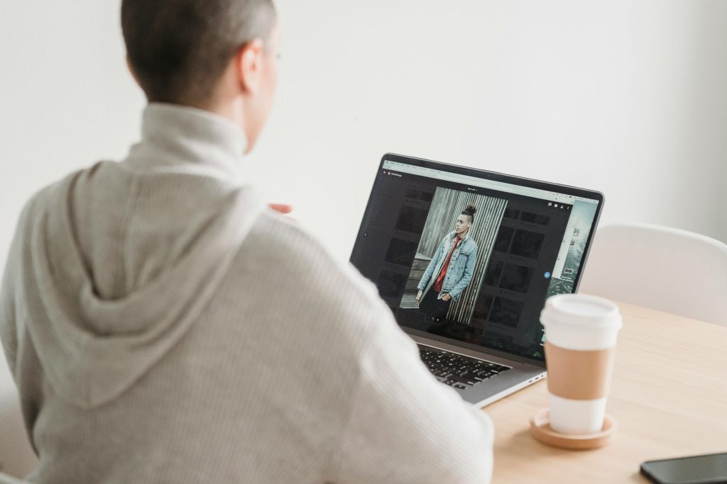 Back view anonymous female in gray hoodie looking at image on netbook screen and sitting at desk with takeaway coffee